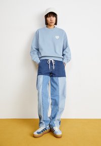 Žena stojící proti bílé stěně, oblečená v bílé kšiltovce, světle modré mikině s detailem srdce, džínech s patchwork vzorem a teniskách.