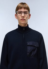 Marinblå stickad zip-up jacka med hög krage, texturerat tyg och en kontrasterande bröstficka i ett glänsande material. Klara bågar på glasögonen.