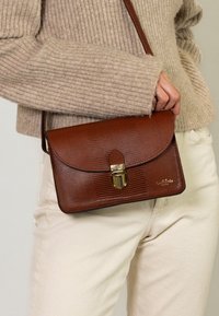 Una persona che indossa un maglione beige e pantaloni crema tiene una borsa a tracolla in pelle marrone testurizzata con chiusura dorata in vita.