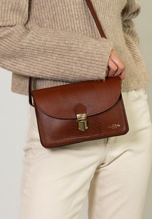 Eine Person trägt einen beigen Pullover und cremefarbene Hose und hält eine strukturiert braune Leder-Crossbody-Tasche mit goldenem Verschluss an der Taille.