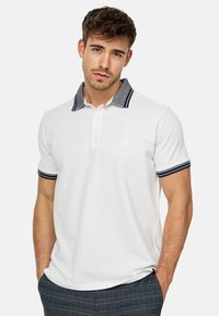Hvid polo-skjorte med en kontrasterende krave i denimstil og marineblå striber. Har en to-knaps knaplukning og et diskret logo på brystet.