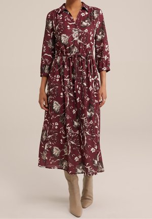 Kvinde iført en burgunderfarvet blomster-midi-kjole med trekvartærmer, knappet forside, samlet talje og beige ankelstøvler.