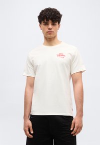 Νεαρός άνδρας με σγουρά μαλλιά που φοράει λευκό μπλουζάκι Levi Strauss & Company και μαύρο παντελόνι, στέκεται μπροστά σε ένα απλό ανοιχτό φόντο.