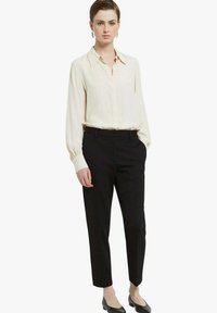 Blusa color crema con maniche lunghe e chiusura con bottoni; abbinata a pantaloni neri su misura. Entrambi i capi presentano una texture liscia e una vestibilità strutturata.