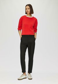 Rode sweatshirt met witte kraagdetails, zwarte taps toelopende broek en beige/witte sneakers met textuuraccenten. Model staat met de handen in de zakken.