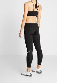 Leggings neri con vita alta, texture liscia e dettaglio del logo bianco, abbinati a un reggiseno sportivo nero e sneakers bianche con motivo.