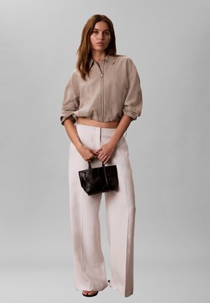 Nő áll, bézs színű rövid cipzáras dzsekit, széles szárú világos rózsaszín nadrágot visel, kezében egy kis fekete, steppelt táska fogantyúkkal.