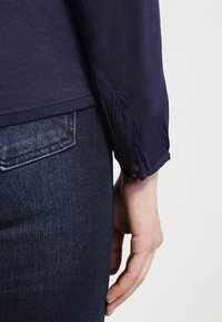 Main droite à côté de la poche d'un jean denim bleu foncé, portant une chemise bleu marine à manches longues avec poignets boutonnés, sur un fond clair.