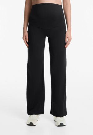 Pantaloni neri a gamba larga con una vita alta e elasticizzata. Tessuto liscio e leggero. Abbinati a scarpe da ginnastica di colore chiaro.
