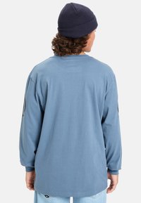 Camisa de manga larga azul de algodón, con cuello redondo y un corte relajado. Un gorro añade un toque casual al cabello rizado.
