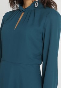 Teal mekko, jossa on avainreiän kaula-aukko, pleated-yksityiskohtia kauluksessa ja pitkät hihat. Luja kangas, jolla on soljuva muotoilu.