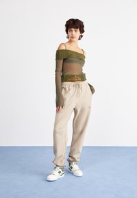 Olivově zelený off-the-shoulder síťovaný top s texturovanými rukávy, v kombinaci s béžovými joggery a bílými teniskami s zelenými akcenty.