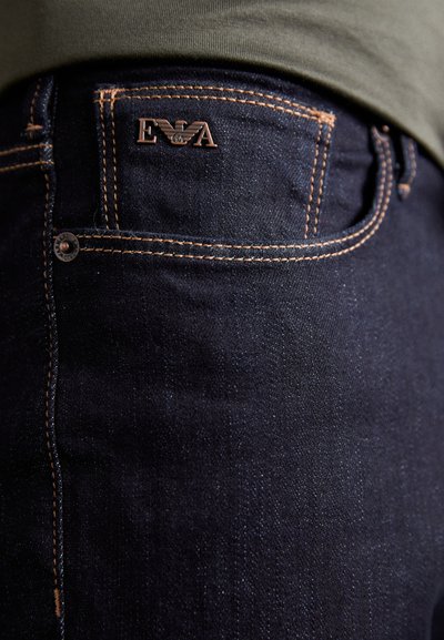 Jean en denim noir avec des surpiqûres orange contrastantes, un petit logo embossé sur la poche arrière, et un design classique à cinq poches.