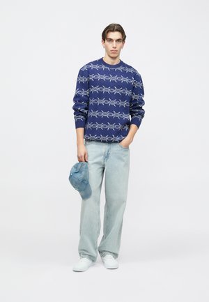 Maglione blu con motivo grafico bianco, jeans di denim chiaro e cappellino blu in mano. Scarpe da ginnastica bianche completano l'outfit casual.