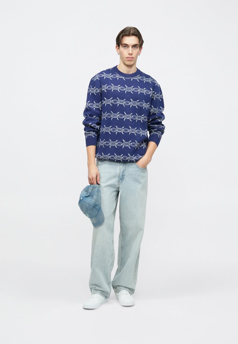 Μπλε πουλόβερ με λευκό γραφικό σχέδιο, ανοιχτόχρωμα τζιν και μπλε καπέλο στο χέρι. Λευκά αθλητικά παπούτσια ολοκληρώνουν την καθημερινή εμφάνιση.