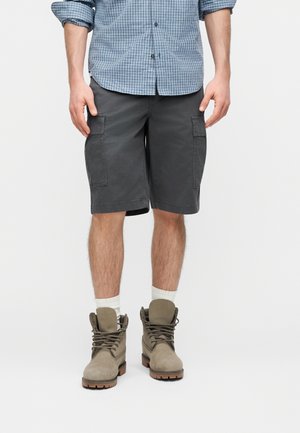 Bărbat purtând pantaloni scurți cargo gri, șosete albe până la gleznă, cizme șnuruite bej și o cămașă albastră cu carouri, cu mânecile lungi suflecate.