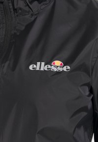 Svart vindjacka med dragkedja och en liten grå "ellesse"-logotyp, prydd med en färgglad emblem ovanför logotypen. Slät textur.