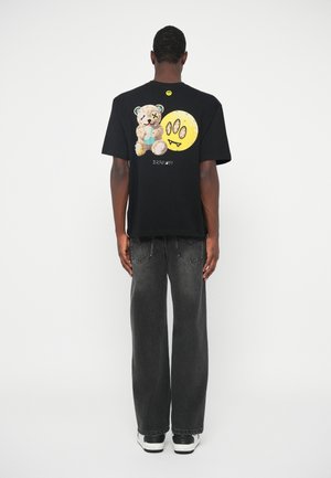Black t-shirt with bear and smiley face graphic on the back, worn with gray wide-leg jeans and black and white sneakers.