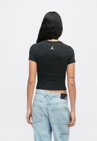 Γυναίκα με μακριά σκούρα μαλλιά, φορώντας ένα μαύρο κοντομάνικο fitted τοπ με άσπρο λογότυπο στην πάνω πλάτη και ανοιχτό μπλε τζιν με μεγάλες τσέπες.