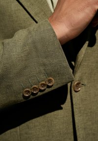 Ärmel eines olivgrünen Blazers aus strukturiertem Stoff, mit fünf hellbraunen Knöpfen und kontrastierendem grünen Stich entlang des Bündchens.