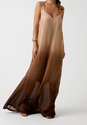 Femme portant une robe longue ombrée marron sans manches avec des bretelles fines et un bracelet doré, debout devant un fond uni.