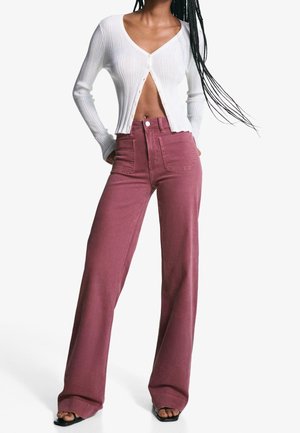 Kobieta w białym krótkim prążkowanym kardiganie oraz wąskich, szerokich, wysokich na talię spodniach w kolorze mauve z kieszeniami z przodu i czarnych sandałach na obcasie.