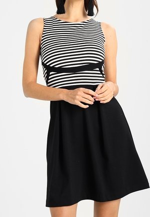 Kvinde iført ærmeløs kjole med sort-hvidt horisontalt stribet top og ensfarvet sort nederdel, hænder samlet ved taljen.