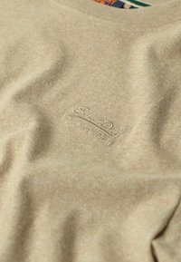 Tessuto beige con il logo "SuperDry" ricamato sottile e caratteri giapponesi in filo abbinato, mostrando la trama e lievi pieghe.