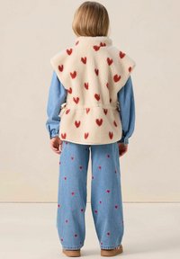 Gilet en polaire crème avec des motifs de cœurs rouges, associé à un jean bleu clair parsemé de cœurs rouges. Cordon de serrage à la taille réglable.