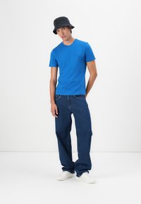 Blauwe katoenen T-shirt gecombineerd met losse, donkerblauwe denim jeans en witte sneakers. Een zwarte bucket hat maakt de outfit compleet.