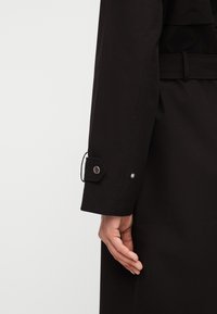 Posnetek roke osebe blizu manšete črne plašče s štirimi gumbi in zanko za pas na enobarvnem ozadju.