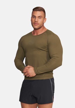 Męźczyzna o muskulaturze, ubrany w oliwkową dopasowaną koszulę z długim rękawem i czarne sportowe spodenki, stoi z rękami splecionymi na jednolitym tle.