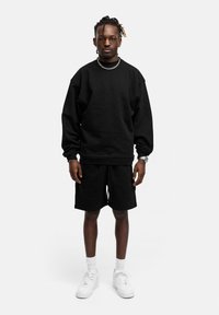 Schwarzer, übergroßer Sweatshirt mit langen Ärmeln, kombiniert mit schwarzen Shorts. Weiße Turnschuhe und eine silberne Kette akzentuieren das Outfit. Einfaches Design.