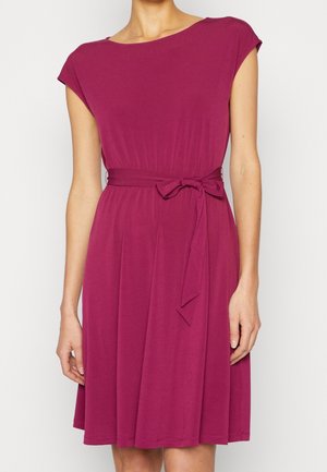 Femme portant une robe magenta mi-longue à manches courtes avec une ceinture nouée à la taille, debout devant un fond blanc uni.