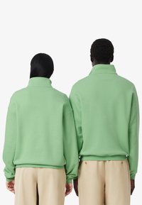 Sudaderas verdes de gran tamaño con cuellos altos, que cuentan con mangas largas y puños acanalados, combinadas con pantalones beige claros.