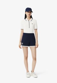 Vitt polotröja med marinblå detaljer, marinblå shorts med sidostripes och en emblem, tillsammans med en marinblå keps och vita sneakers.