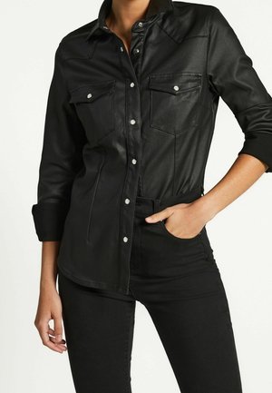 Femme portant une chemise noire en similicuir boutonnée avec des poches à rabat sur la poitrine et un pantalon noir, une main dans la poche et les manches retroussées.