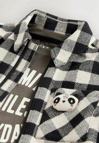 Veste en flanelle à carreaux noir et blanc avec une texture confortable, ornée d'une appliqué de panda et d'un T-shirt gris en dessous affichant un texte blanc.