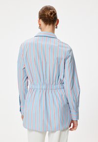 Camisa de botones a rayas en azul y blanco con acentos rojos, cintura cinchada, mangas largas y cuello para un ajuste estructurado.