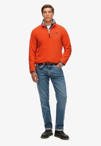 Pulóver de forro polar naranja con cuello de media cremallera, superpuesto a una camisa de cuadros azul, combinado con jeans de un azul claro y zapatos negros.
