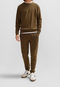 Felpa verde oliva con polsini e orlo a costine, abbinata a pantaloni jogger coordinati. Camicia bianca a strati sotto, e sneakers con dettagli marroni.