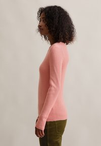 Frau mit lockigem Haar, die einen eng anliegenden, rosa gerippten Pullover und grüne Cordhosen trägt und seitlich vor einem schlichten Hintergrund steht.