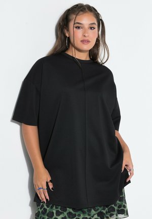 Ung kvinde med langt brunt hår og øreringe i hoops har en oversize sort t-shirt og et grønt leopardmønstret skørt på og poserer mod en hvid væg.