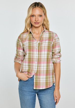 Kobieta ubrana w różowo-białą i oliwkową koszulę w kratę z podwiniętymi rękawami oraz niebieskie dżinsy, stojąca z jedną ręką w kieszeni.