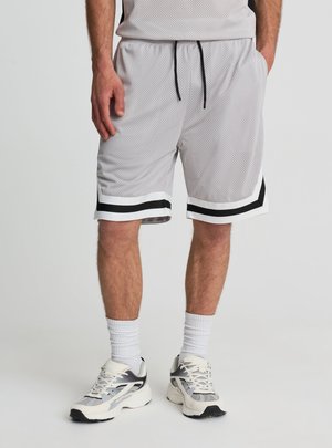 Uomo che indossa pantaloncini da basket in mesh grigio chiaro con rifiniture nere e bianche, calzini bianchi e scarpe da ginnastica bianche e grigie.