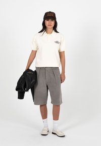 Femme debout, portant une casquette marron, un polo crème, un short gris jusqu'aux genoux, des chaussettes blanches et des mocassins crème, tenant une veste en cuir noire.