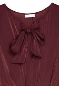 Blouse bordeaux avec une finition texturée, présentant un nœud au cou et une ouverture en forme de V. Tissu lisse avec un léger éclat et des détails de drapés.