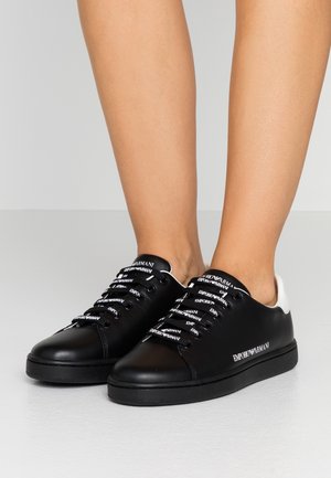 Czarne niskie sneakersy z białymi logo "Emporio Armani" na sznurowadłach, języku i boku, noszone na bose stopy na neutralnym tle.