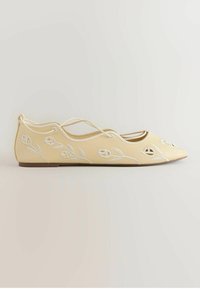 Bailarina plana color crema con bordado floral blanco, bordes en forma de ondas y delgadas correas cruzadas en el tobillo, exhibida sobre un fondo liso.