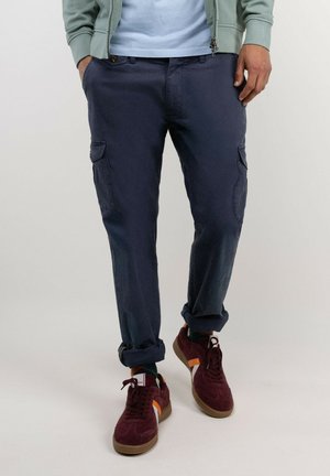 Hombre con pantalones cargo azul marino con los bajos remangados, zapatillas burdeos con rayas blancas y naranjas, camisa azul claro y sudadera verde con cremallera.